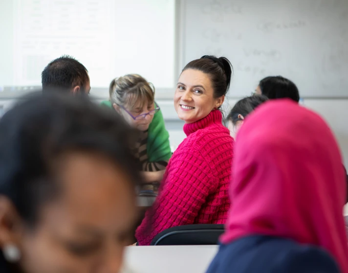 A woman in a bright pink sweater smiles at the camera while sitting in a classroom with other students working in the background. A woman in a bright pink sweater smiles at the camera while sitting in a classroom with other students working in the background.